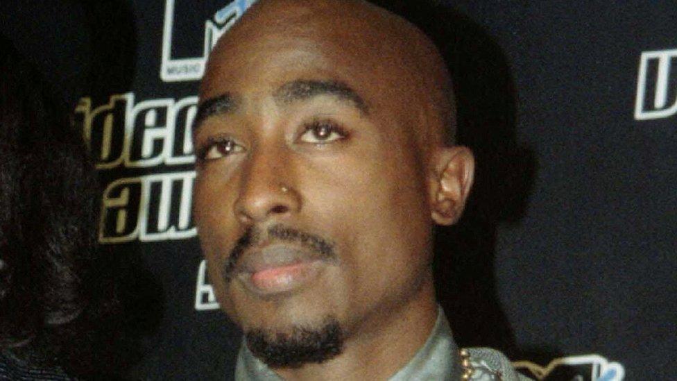 A close-up of Tupac Shakur - a black man with bald head and goatee beard. He has a gold stud in his left nostrial. He appears to be standing in front of a hoarding on a red carpet at some sort of event. Camera lights can be seen reflected in his eyes.