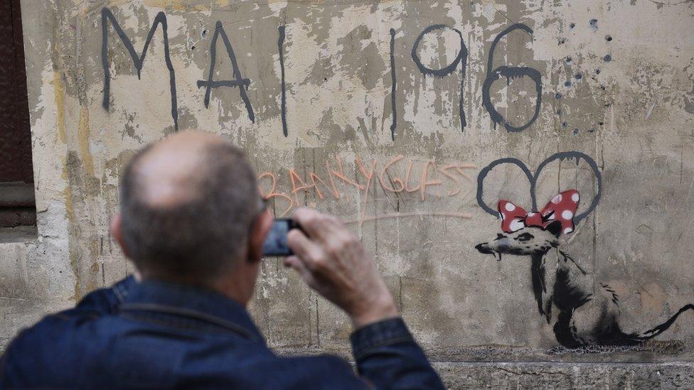 A man takes photographs of a recent artwork believed to be attributed to Banksy showing a rat wearing a Minnie Mouse bow under "May 1968" in the Sorbonne neighbourhood