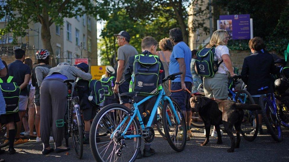 A young boy stands with his bike at the gates to St Martin school, surrounded by parents and children.