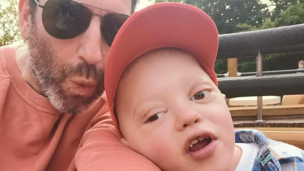 Boy in orange cap leans on his father's arm. Father has a beard and dark glasses.