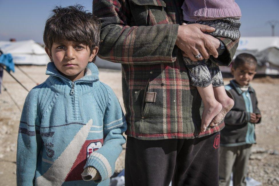 A boy, 7, looks at the camera at a refugee camp in Syria. He stands next to his father, Hisham (not his real name), who carries an infant. We cannot see either of their faces. Another child looks on from behind.