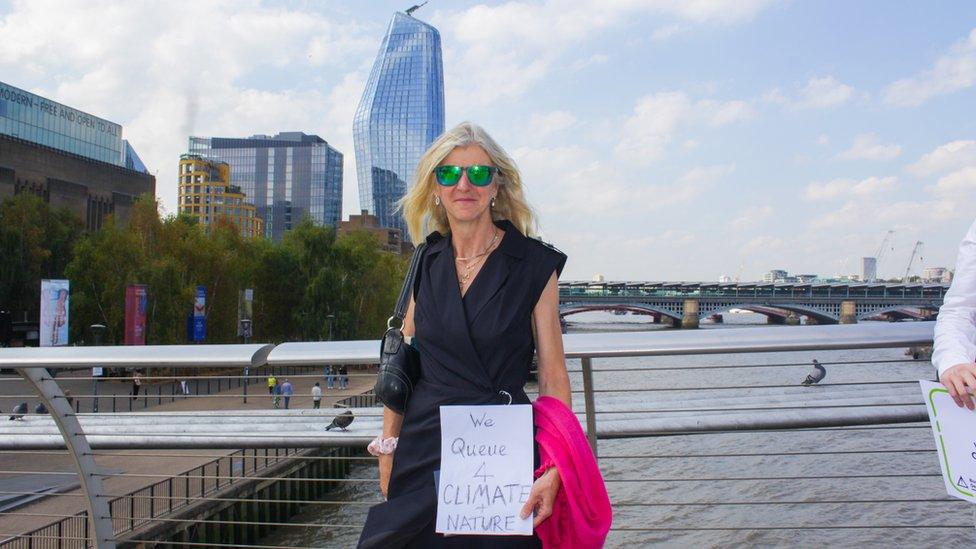 A woman stands on Millennium bridge, holding a sign that says 'we queue for climate and nature'