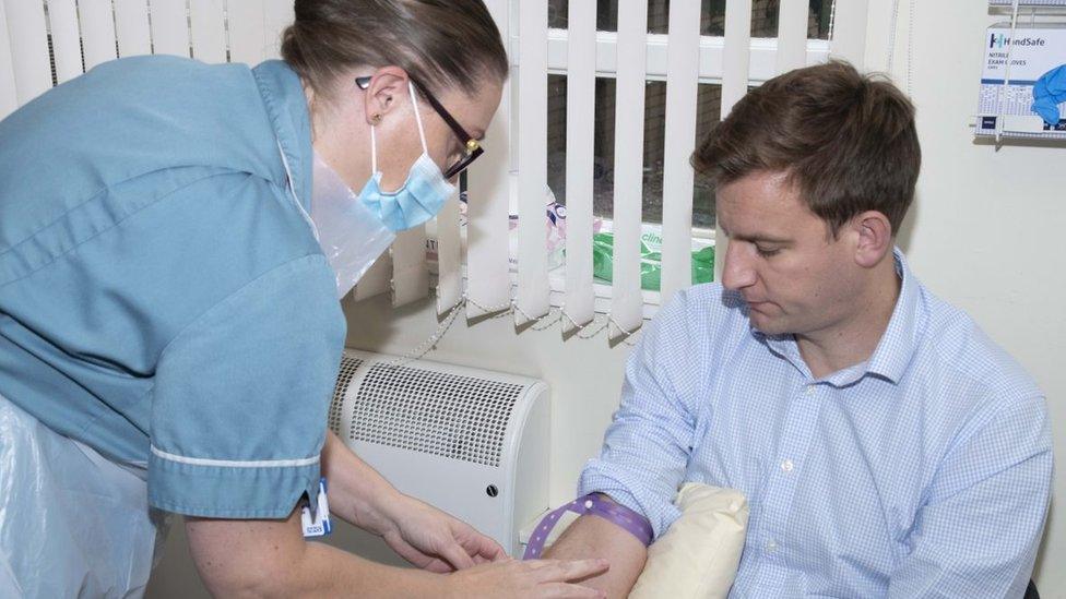 Man with short brown hair has his sleeve rolled up whilst a nurse uses a syringe to take a sample