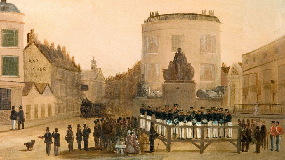 A painting showing a brass band in front of a statue in Weymouth, Dorset, in 1844. The band is raised on a wooden stand and are in white and blue uniforms. A crows of men in stove-pipe hats are watching on the right, accompanied by two women in hooped dresses and two children in white clothes. On the left are more on-lookers, including two red-uniformed soldiers