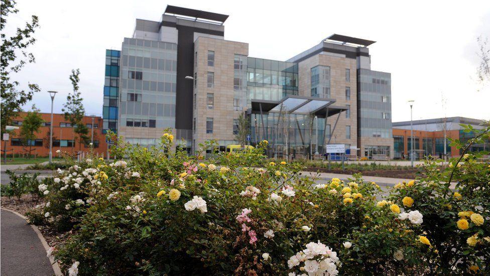 Peterborough City Hospital, a multi-storey modern building with large windows. In front of it is a bed of multi-coloured roses 