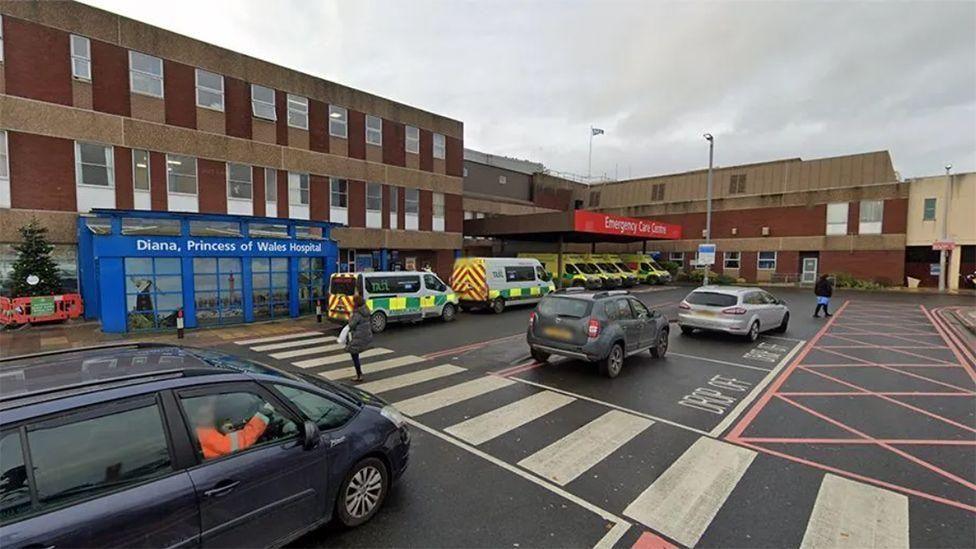 Car drop-off point in front of a low, brown building with a blue sign reading: Diana, Princess of Wales Hospital