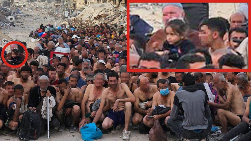 A group of dozens of detained Gazan men pictured in their underwear, during what appears to be an Israeli check for weapons and signs of any links to Hamas. They are sitting or squatting, some young and some older. At the far left of the picture, in the middle of the group, a little girl with dark hair can be seen. The BBC has chosen to blur the faces of the men nearer the front of the picture. In the background is a destroyed building, collapsed into rubble.   