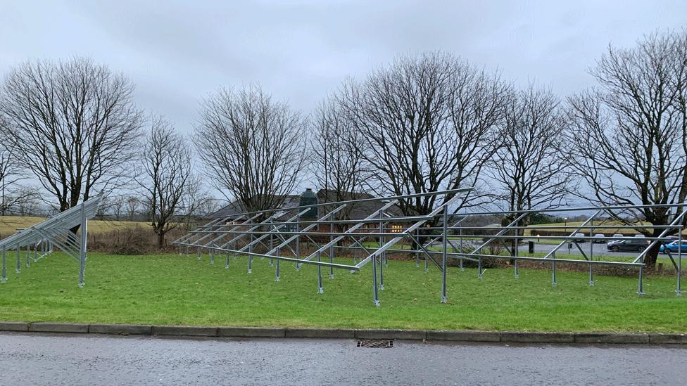 The metal frames for solar panels sit on top of grass with bare trees in the background and a car park behind them