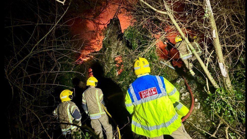 Backs of four firefighters with stone building in the background - it is engulfed by fire that is giving off a bright orange glow.