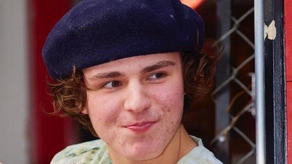 Harvey Owen has curly brown hair and is wearing a navy blue beret style hat. He is wearing a green t-shirt and smiling, while looking to the side.