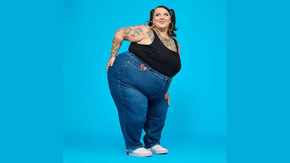 Sophie who is a UK size 24 to 26 model on a shoot wearing blue jeans and a black vest top and glamorous make up, posing with her hand on one hip against a bright blue background. she has tattoos on her arm and chest.