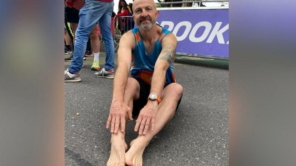 John Hillage sat on the ground with his arms stretched towards his bare feet. He has a grey beard and is wearing sports gear.