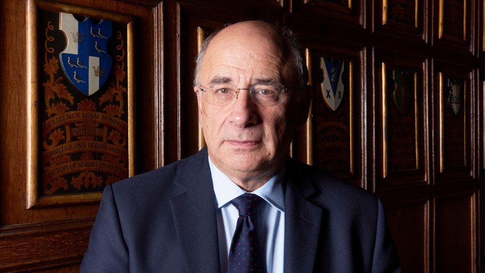 A head-and-shoulders portrait of Sir Brian Leveson, a white man with rimless glasses and grey hair at the sides of his head, wearing a navy suit with a light blue shirt and navy tie. He is standing in a wood-panelled room with heraldic designs behind him and is light by soft light coming from the left of the picture.