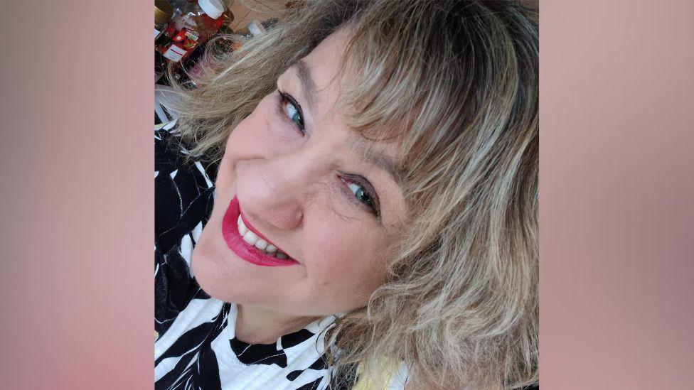 A close-up selfie of a woman with blonde hair and bright pink lipstick smiling at the camera. The woman is wearing a black and white top. She appears to be standing in a kitchen and there is a bottle of ketchup behind her.