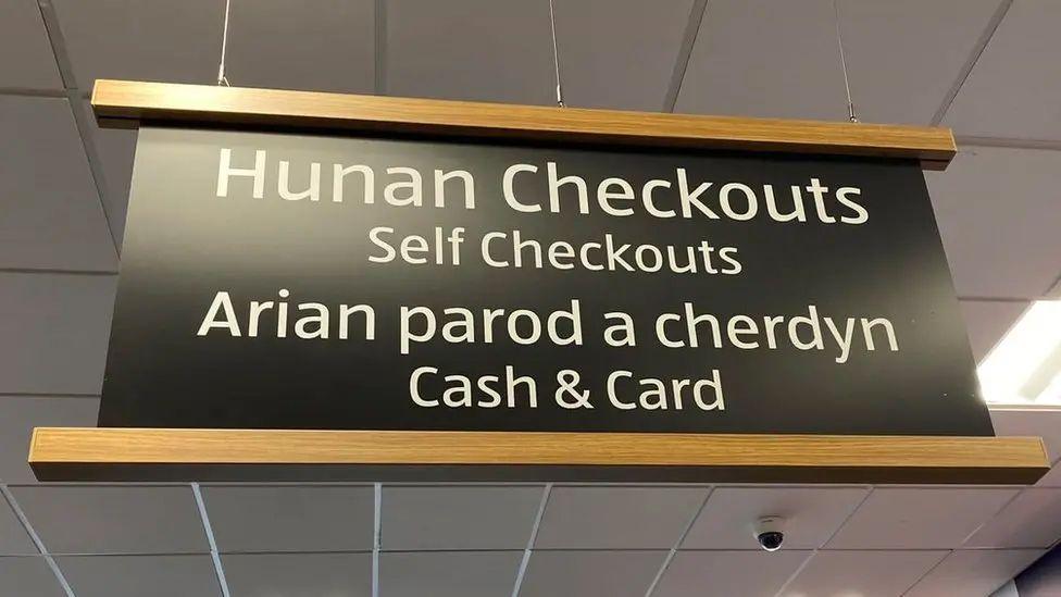 A black sign hanging from the ceiling of an Aldi supermarket reads: "Hunan Checkouts/ Self Checkouts. Arian parod a cherdyn/ Cash & Card."