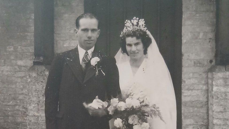 A sepia toned black and white photo showing Wally Marshall and Vera Edwards on their wedding day, with a brick built church with a large door behind them. Wally has slicked-back short hair and is clean shaven. He is wearing a dark suit over a white shirt and tie, with a carnation button hole. He is holding hands with Vera, who is in a white v-necked dress with a white veil and a tiara-style headress on her short curling hair. She is carrying a bouquet. They are both starting to smile. 
