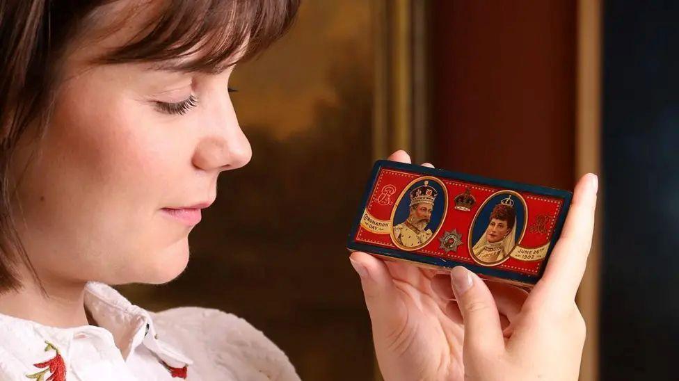 An auctioneer holds the silver chocolate tin.