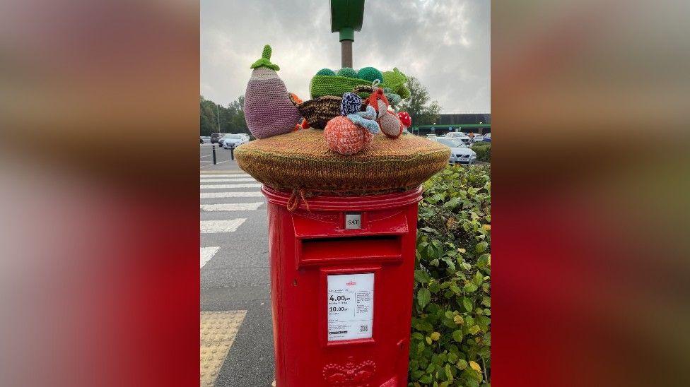 A photo of a red post box with a knitted topper featuring different fruit and veg.