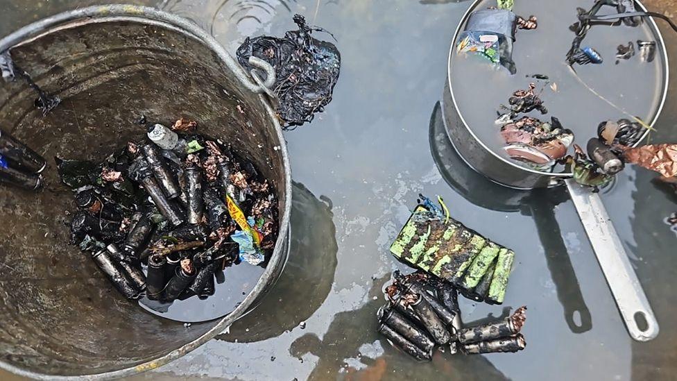 Aftermath of the fire with a metal bucket and saucepan filled with charred batteries and surrounded by water 