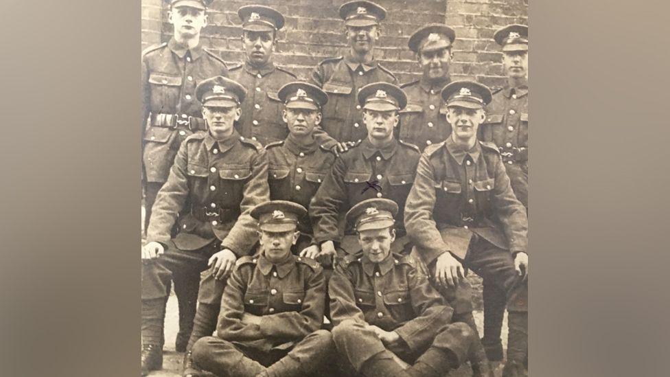 A sepia photograph of 11 young male soldiers dressed in uniform posing for the cameras in three rows with all looking at the camera.