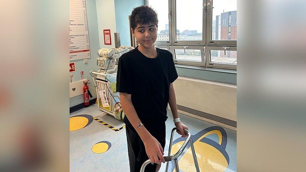 Zak is standing in a hospital room with his hands on a walking aid. 