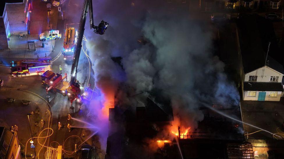 A drone shot of flames and large plumes of smoke coming from a building. There are large amounts of emergency service vehicles in the road next to the building.