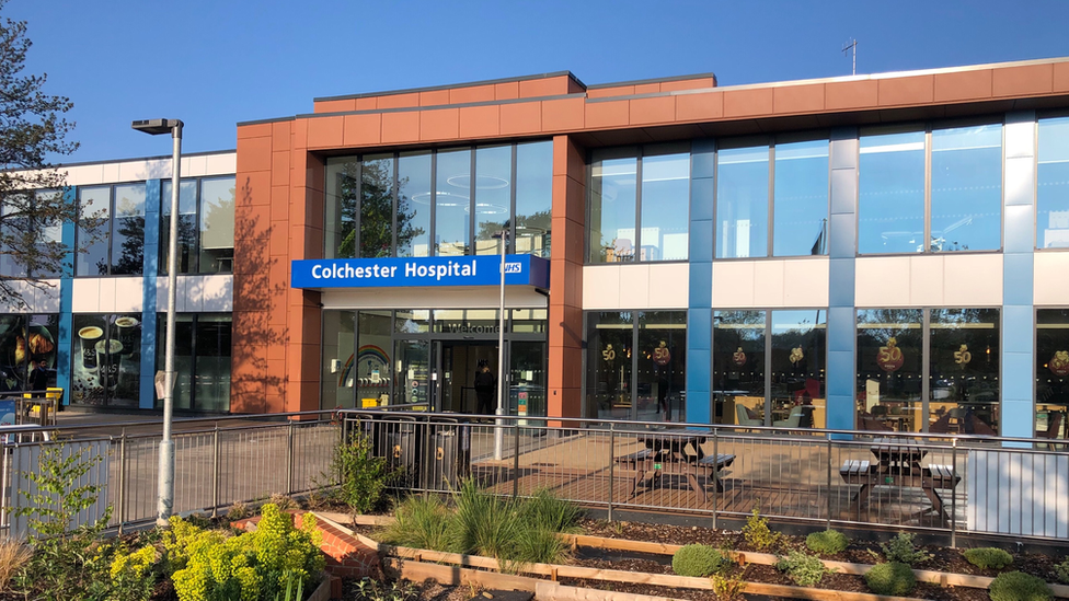 The entrance of Colchester Hospital, which is a two-storey building with a facade mostly covered in glass windows. It is a mixture of orange, blue and white panels and there is a sign that reads "Colchester Hospital" above the entrance. In front of the building is pavement, benches and some plants.