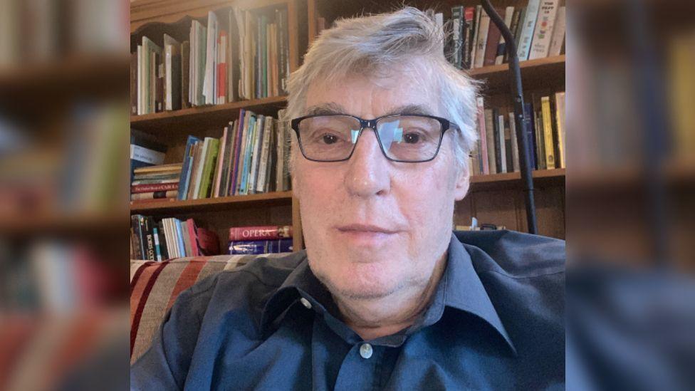 Jerry Cowhig sitting on a striped sofa in front of a wooden bookcase. There are lots of thin books and documents stacked vertically on the shelves, and a black standing lamp lit above Mr Cowhig's head. He is wearing a blue button up shirt and black framed glasses. He has short white hair that is flipped over to the left side. 