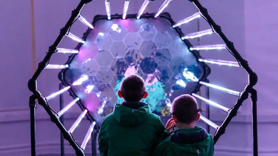 Two boys stand facing a purple hexagonal light installation made up of long tubes on the sides and lots of smaller hexagons in the middle that shimmer as though in water