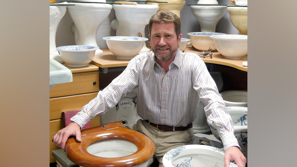 A man pictured kneeling on floor with both hands on two antique loos

The man has brown hair and a beard, wearing beige chinos and a stripy shirt with a brown belt.