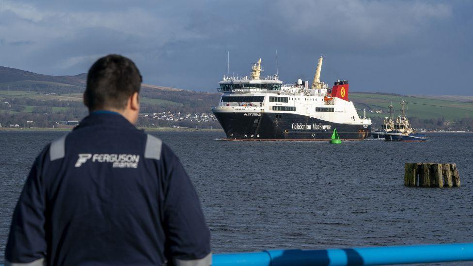 A Ferguson Marine worker, wearing company overalls, stands in the foreground looking out over the River Clyde towards a CalMac ship 