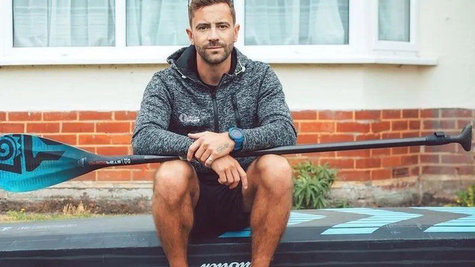 Man in shorts sat on a paddleboard with a oar across his lap a brick building can be seen behind him.