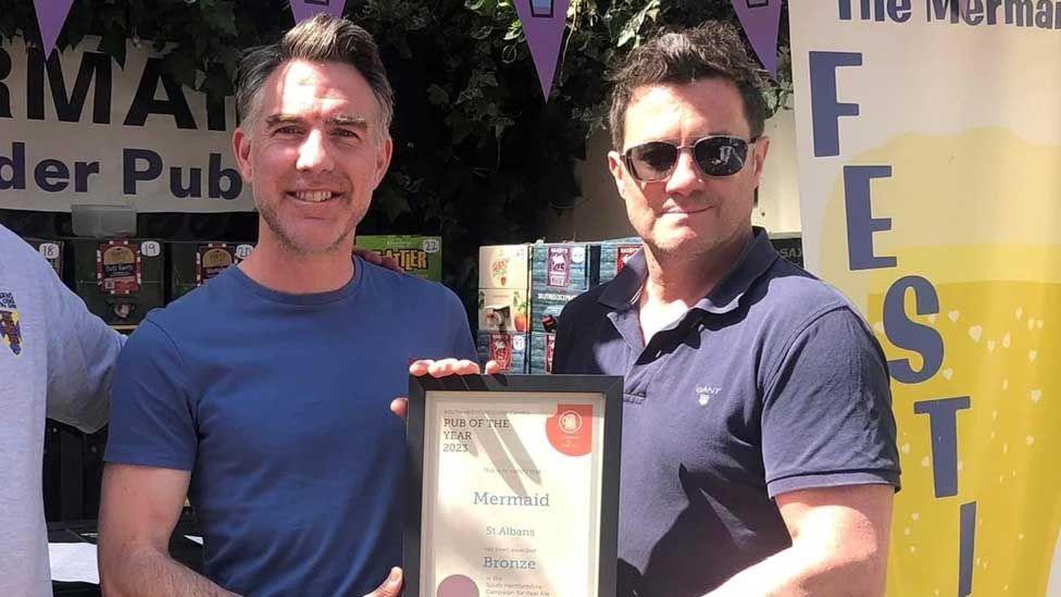 John Cusworth [left] in a royal blue T-shirt and Mark Powell [right] in a navy blue T-shirt holding a black-framed certificate for a bronze award in the South Herts Campaign for Real Ale Pub of the Year competition 2023 