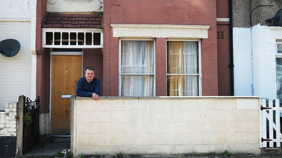Roye Chapman is leaning on a low wall in front of a red brick house with two windows covered by lace curtains and a wooden door. The house has a small front yard with some grass and plants. Roye is wearing a blue long-sleeve shirt