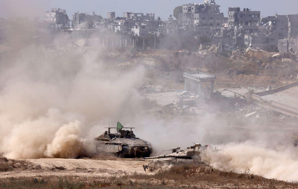 An Israeli tank and an armoured personnel carrier inside the Gaza Strip near the Israel-Gaza border, June 2024 - damaged buildings are visible in the background
 