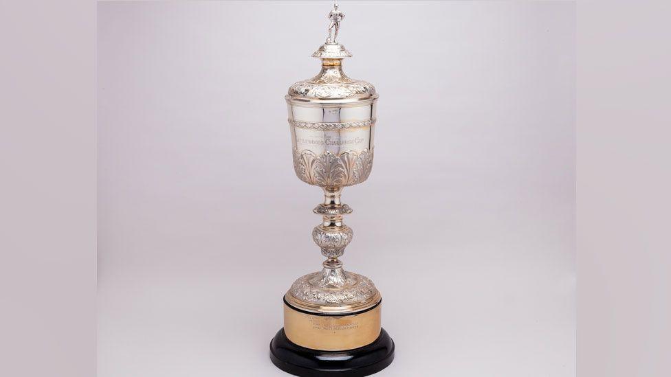 The Littlewoods League Cup trophy, a solid silver chalice-like cup. It has a lid on which stands the figure of a male footballer with his hands on his hips and his left leg resting on a ball. The cup is ornate with a leaf pattern around its stem and base. It is resting on a gilded base, which in turn is resting on a wooden base.