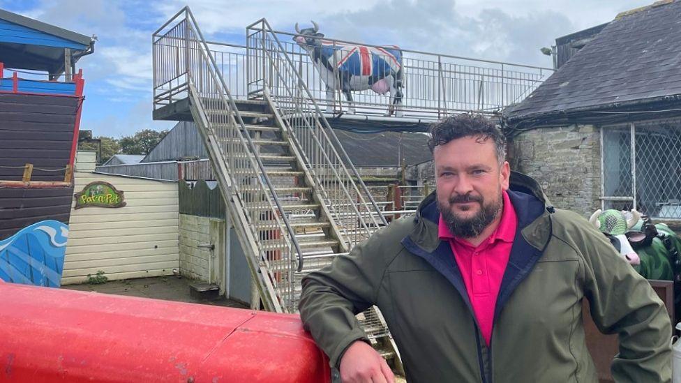 Diaryland general manager Keith Thompson, with dark brown curly hair, a beard, wearing a pink polo short under a green outdoor coat. He is leaning against a tractor