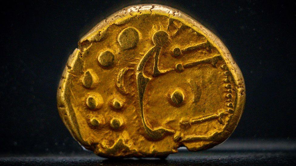 A close-up of a gold Iron Age coin. It is a rounded oval in shape and has dots and shapes struck on it.