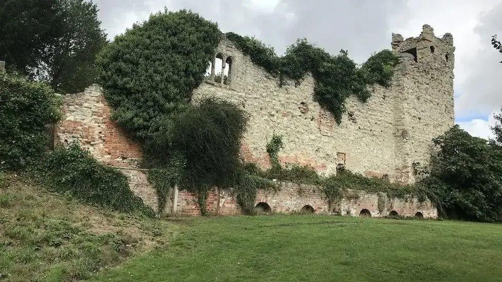 A large ruined castle wall sits on a nearly mown field, with huge amounts of Ivy growing out of its brickwork. The remains of a watchtower are visible at one end.