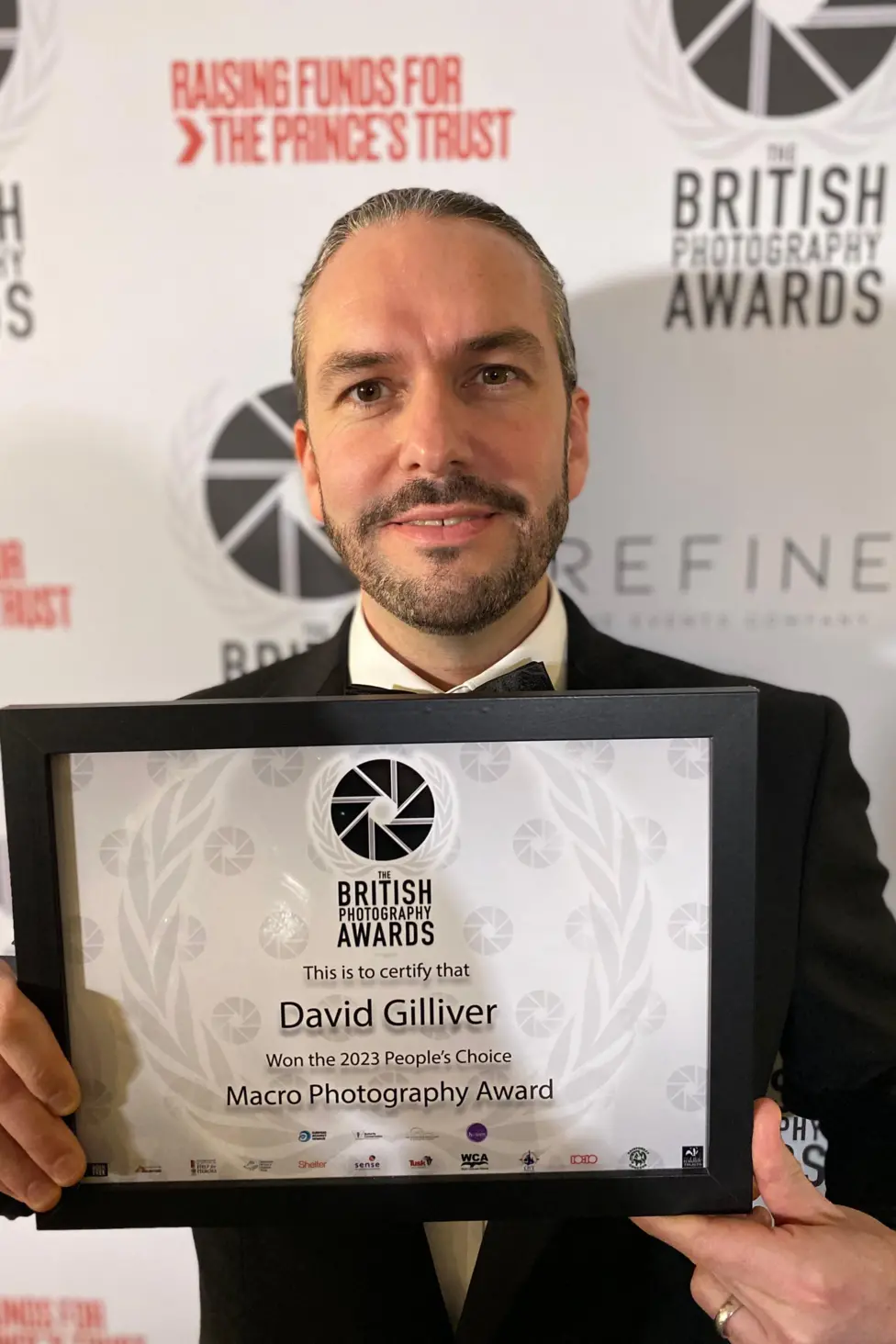 Mr Gilliver looks directly at the camera, holding his framed award certificate. He is wearing a black suit jacket and tie. He has a trimmed beard and hair smartly slicked back.