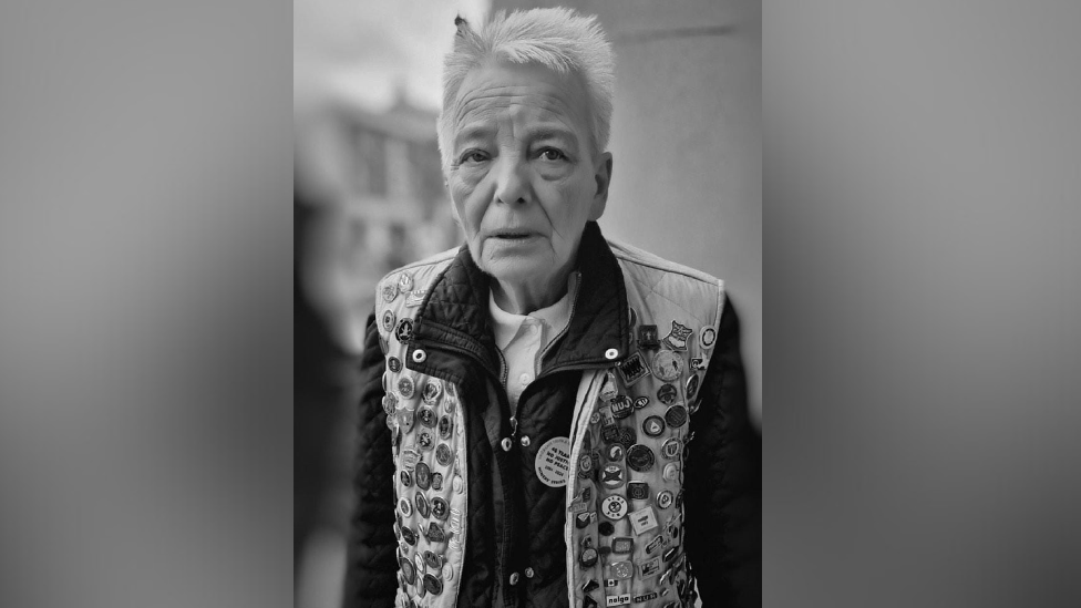 A woman in her 70s with short white hair and jacket covered in badges. She is looking into the camera.