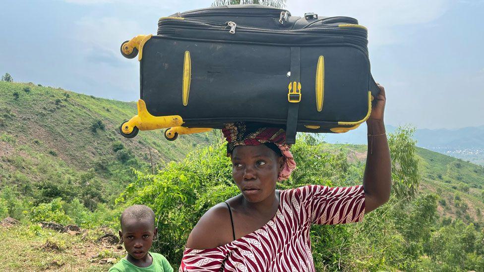 A woman with a black and yellow suitcase on her head and a child are pictured on a hill in Burundi after crossing over from DR Congo