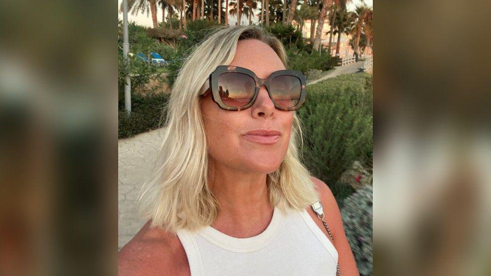 Selfie of Becky Curtis, wearing a white vest top and large, brown sunglasses, while on holiday with palm trees in the background