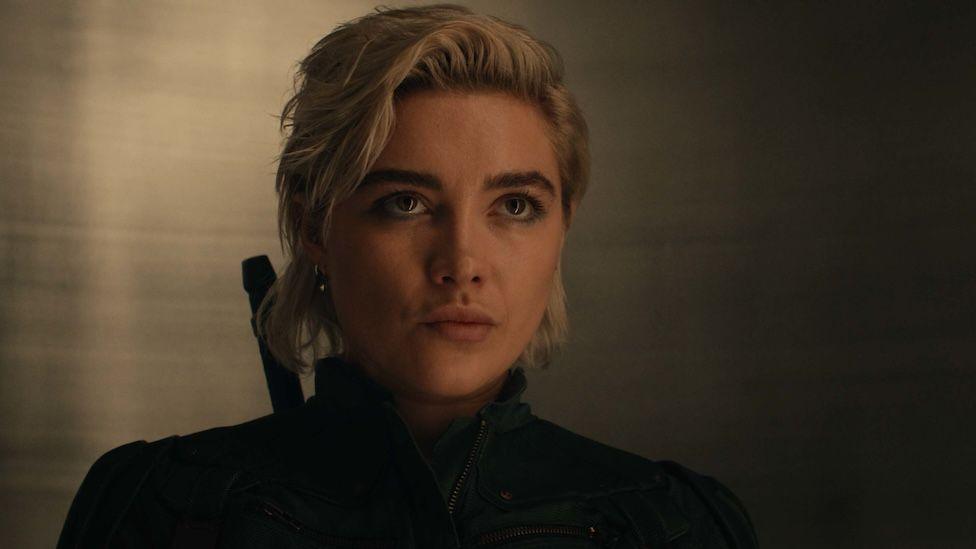 Florence Pugh appears with short blonde hair and wears a dark outfit.