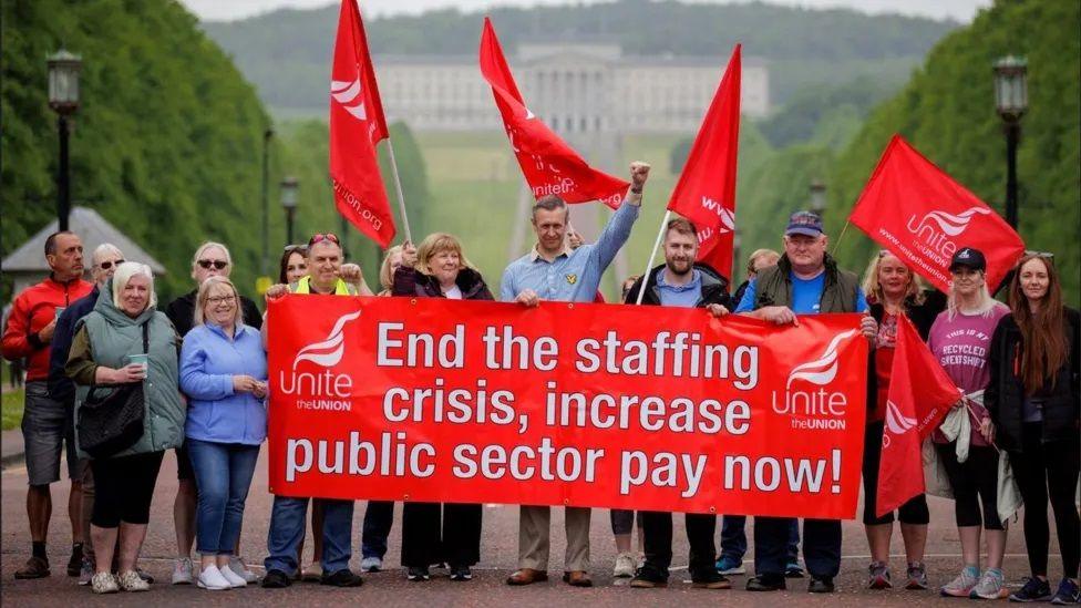 School support staff striking outside Stormont, holding red unite union flags and a banner which reads: "end the staffing crisi, increase public sector pay now!"