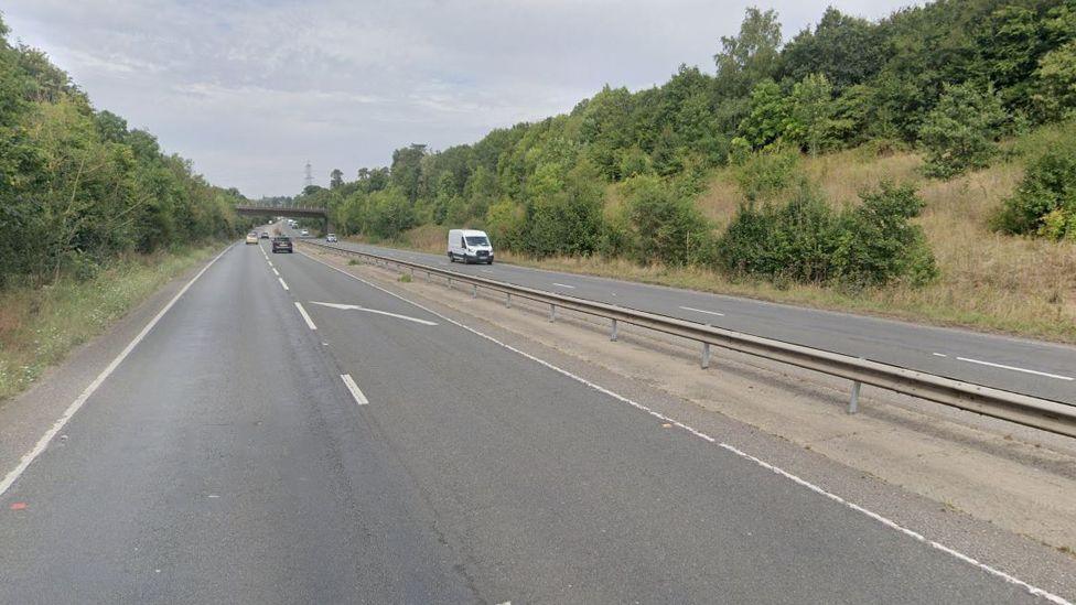 A602 where it changes from a dual carriageway to a single lane carriageway, with a white arrow marking guiding drivers into the single lane. There is a central reservation in the middle. There are banks either side with trees and a bridge over the road in the distance. A few cars are seen in the distance.