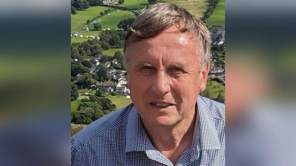 Phil Johnston smiles at the camera. He has short grey and dark hair and wears a checked shirt. There is a view of Coniston village behind him, which shows the countryside.