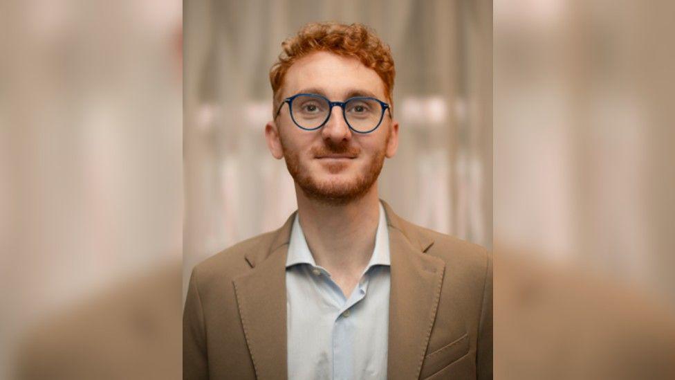 A close-up headshot of Gaetano smiling to camera. He is a red-head with short, curly hair and a short beard. He is wearing a beige jacket and pale blue open-necked shirt and has wide, blue-rimmed glasses on.