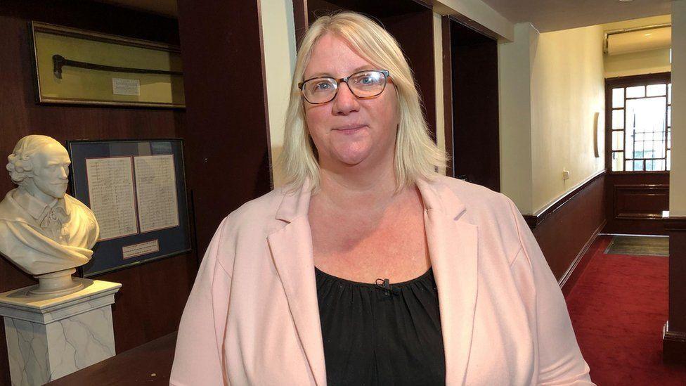 Jo Gordon with long blond hair and glasses wearing a pink cardigan and black top in a red-carpeted foyer area. A stone bust of a man's head sits on an adjacent shelf.