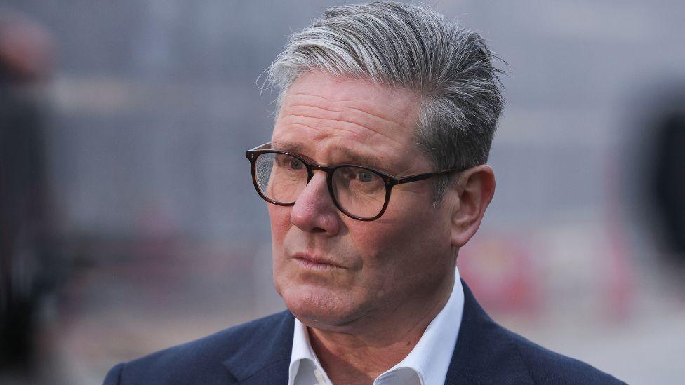 Keir Starmer looks off to the side of the shot - he has grey hair and glasses and is wearing a dark suit and white shirt